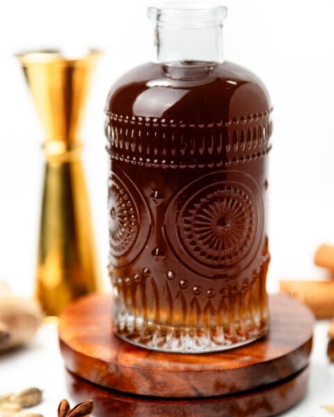 A decorative glass bottle filled with dark liquid sitting on a wooden coaster, surrounded by spices.