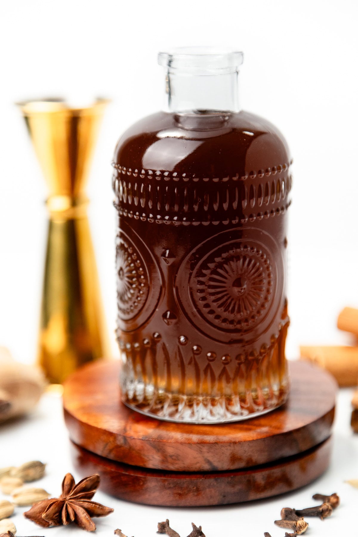 A decorative glass bottle filled with dark liquid sitting on a wooden coaster, surrounded by spices.