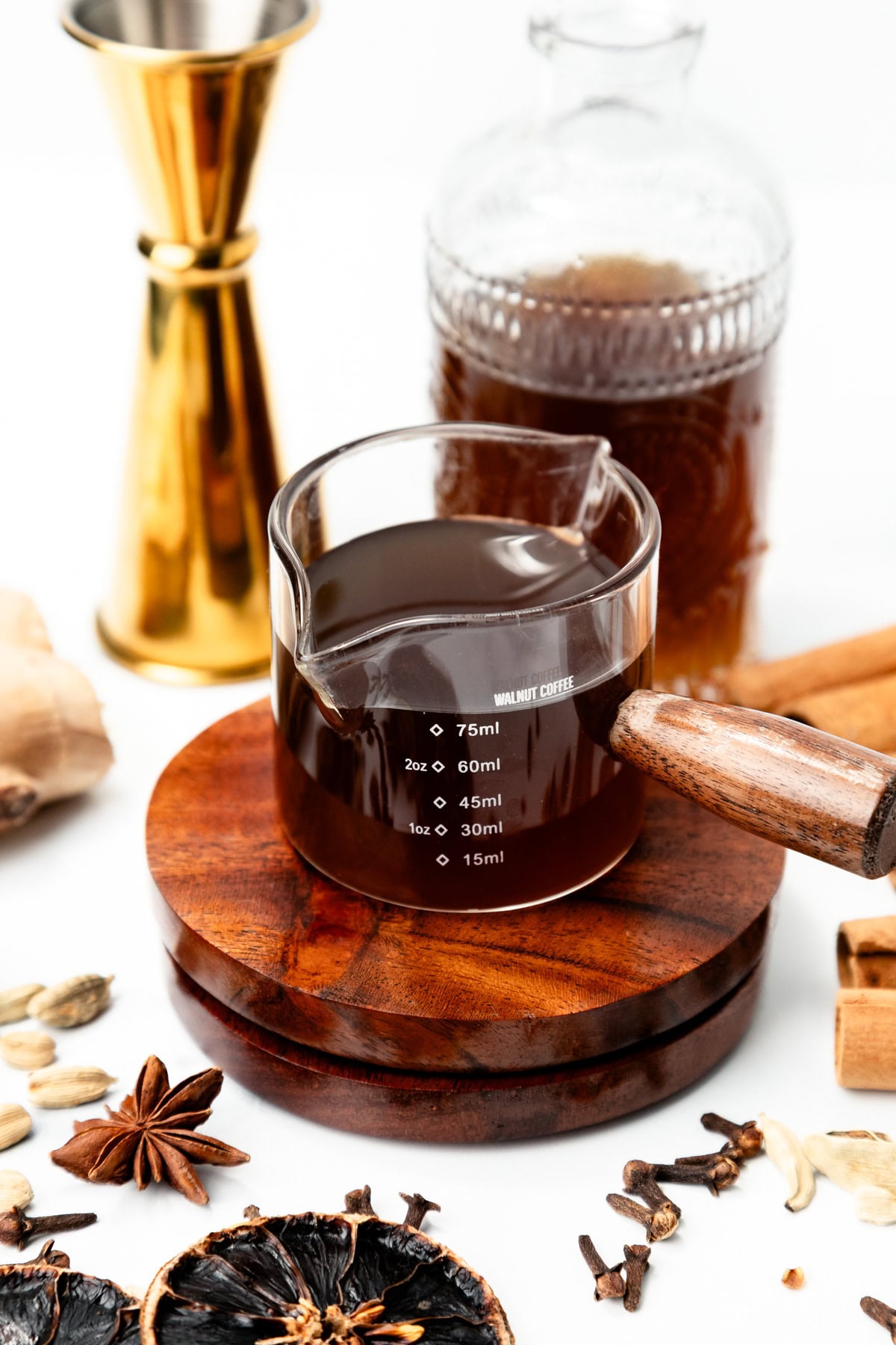 A glass measuring cup filled with dark coffee, resting on a wooden coaster surrounded by whole spices.