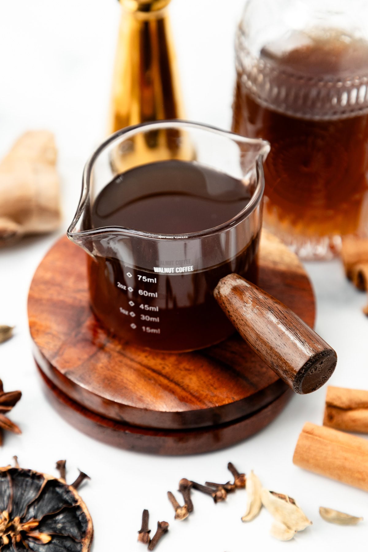 A glass measuring beaker filled with coffee, resting on a wooden coaster surrounded by spices.