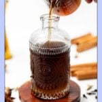 Dark spiced syrup being poured into a decorative glass bottle on a wooden coaster.
