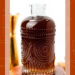 A glass bottle of dark spiced simple syrup on a wooden coaster with star anise and cloves.