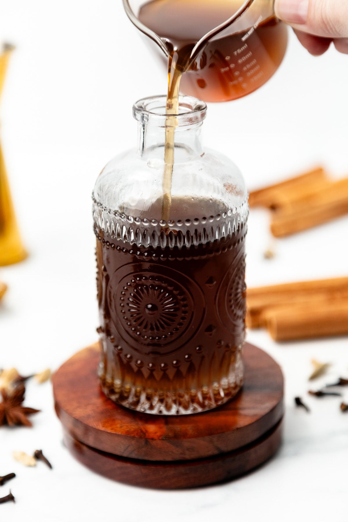 Liquid being poured from a glass measuring pitcher into an embossed glass bottle on a wooden coaster.